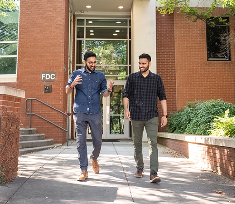 Two men walk out of a brick academic building, smiling and gesturing with their hands.