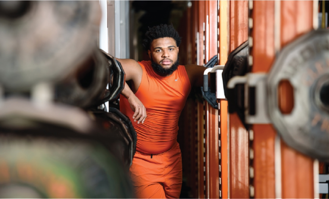 Portrait of a Clemson football player leaning on weightlifting equipment in all-orange practice wear. 