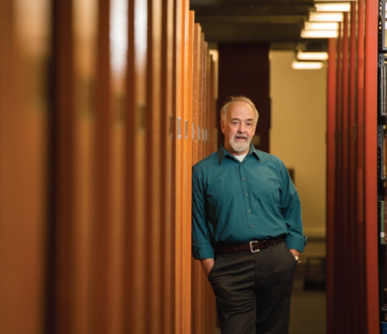 Portrait of a man wearing business casual dress, leaning against book shelves in the library, with hands in his pockets. 