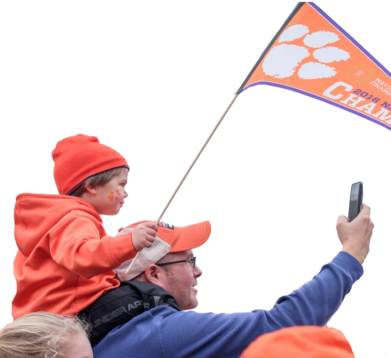 A man holding up his phone to take a picture while a child rides on his shoulders and holds a Clemson branded flag.