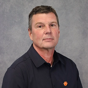 Professional headshot of a man wearing a dark shirt with an orange logo, against a gray background.