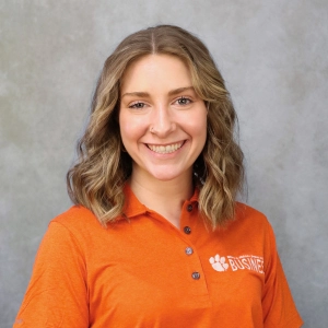 Smiling woman wearing an orange polo shirt with "BUSINESS" printed on it, set against a gray background.