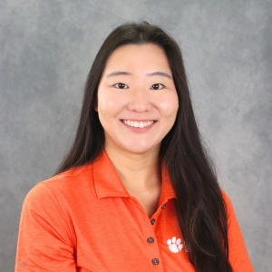 Professional headshot of a woman in an orange polo shirt with a Clemson University logo.