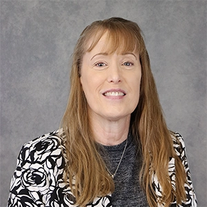 Professional headshot of a woman with long hair wearing a floral blazer against a gray background.