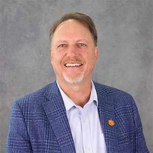 Professional headshot of a man in a blue blazer, smiling against a gray backdrop.