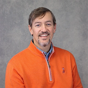 Professional headshot of a smiling man in an orange sweater.