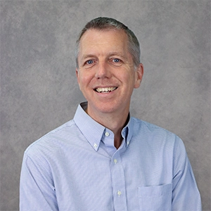 Professional headshot of a smiling man in a light blue button-up shirt.