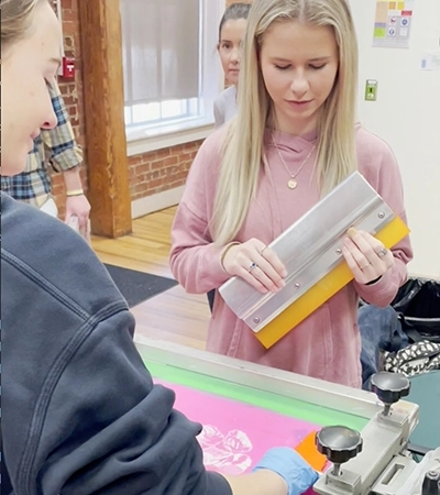 A woman holds a squeegee while observing another person screen printing in an art studio setting.