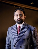 A well-dressed man in a stylish striped suit and red tie poses confidently against a dark background.