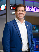 Professional portrait of a smiling man in a blue blazer, standing in a modern office environment.