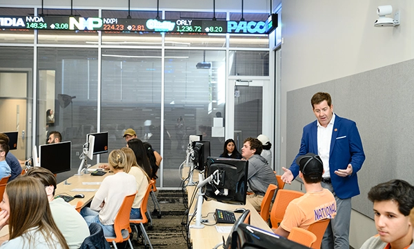 A classroom setting with a speaker presenting to students at computers, featuring a digital display of stock tickers in the background.