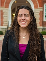 A smiling young woman with long curly hair, dressed in a black blazer and pink top, stands in front of a brick building.