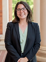 Professional headshot of a smiling woman in a black blazer, standing outdoors with a blurred background.