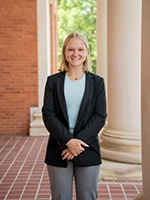 A smiling young woman in a blazer stands confidently in an outdoor setting with brick columns in the background.