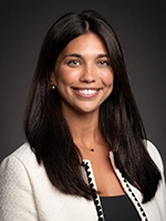 Professional headshot of a woman with long dark hair and a warm smile, wearing a light-colored blazer.