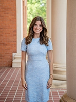 A smiling woman in a light blue dress stands in an outdoor corridor with brick walls and columns.