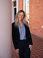 A smiling woman in professional attire stands beside a column in front of a brick building.