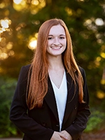 A confident young woman with long red hair smiles while dressed in a black blazer against a natural, softly blurred background.