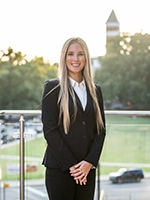 A professional woman in a black suit stands confidently outdoors, smiling with a blurred cityscape in the background.