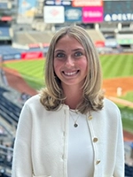 A smiling woman with long, blonde hair stands in a stadium, dressed in a white sweater, with baseball field and stands in the background.