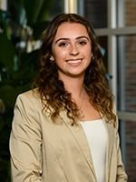 Professional portrait of a smiling young woman with curly hair, wearing a light-colored blazer and standing in a well-lit indoor setting.