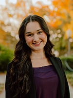 A young woman with long dark hair smiling in a professional outfit, set against a backdrop of autumn foliage.