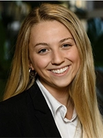 Headshot of a smiling young woman with long blonde hair, wearing a black blazer and white shirt.