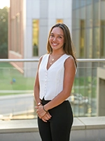 A young woman stands smiling in a professional outfit, with modern architecture in the background.