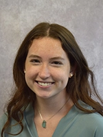 Headshot of a smiling young woman with wavy brown hair, wearing a light blue blouse and a necklace with a green pendant.