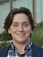 Smiling young man with medium-length brown hair, wearing a plaid shirt and blazer, standing outdoors with greenery in the background.