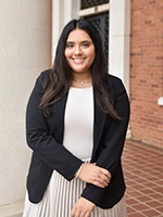 A smiling young woman in a black blazer and white blouse, standing confidently in a professional setting.