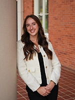 A smiling woman in a stylish white jacket poses confidently against a brick wall, with her arms crossed.