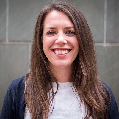 Professional portrait of a smiling woman with long hair, wearing a white top and navy cardigan, against a stone background.