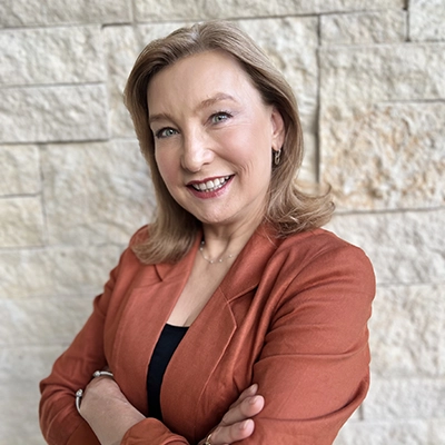 A professional headshot of a woman in an orange blazer, smiling confidently with her arms crossed against a textured stone background.