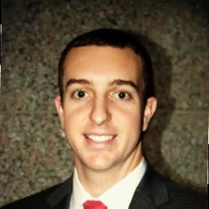 Professional headshot of a smiling man in a suit and tie, set against a stone background.