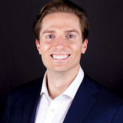 Professional headshot of a smiling man in a dark suit jacket and white shirt against a black background.