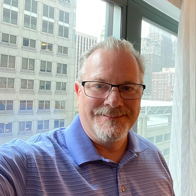 A middle-aged man with glasses and a light blue polo shirt smiling for a selfie in front of a large window with city buildings visible in the background.