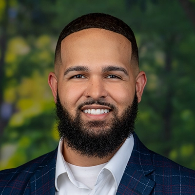 A smiling professional man with a beard, wearing a blue plaid suit and white shirt, set against a blurred green background.