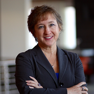 Professional headshot of a woman in a gray blazer, smiling confidently with arms crossed.