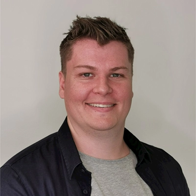 Headshot of a young man with short hair, wearing a navy blue shirt over a gray t-shirt, smiling at the camera.