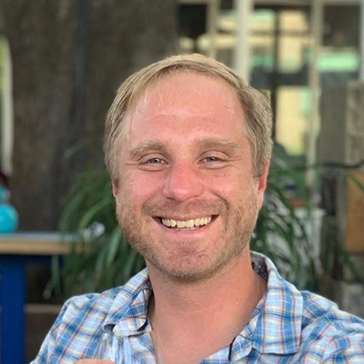 Smiling man with short blond hair wearing a plaid shirt, sitting outdoors in a casual setting.