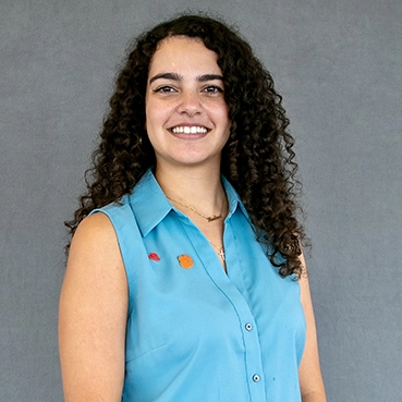 A smiling woman with curly hair wearing a light blue shirt, standing against a gray background.