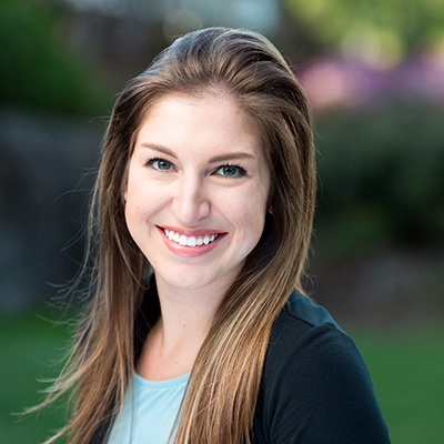 A smiling woman with long brown hair, wearing a black jacket and set against a blurred green background.