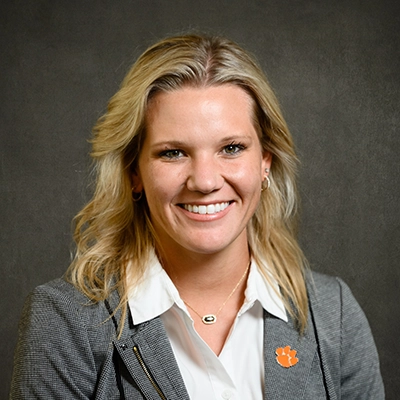 Professional headshot of a woman with blonde hair wearing a gray blazer and white shirt, smiling confidently.