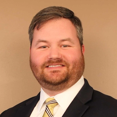 Professional headshot of a man with a beard, wearing a suit and tie, smiling against a neutral background.