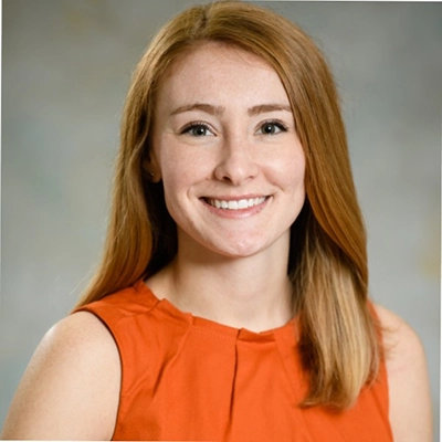 Professional headshot of a woman with long auburn hair wearing an orange sleeveless top.