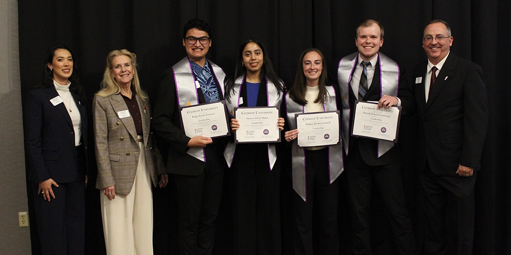 Group of graduates proudly holding their certificates at a graduation ceremony, accompanied by a faculty member.