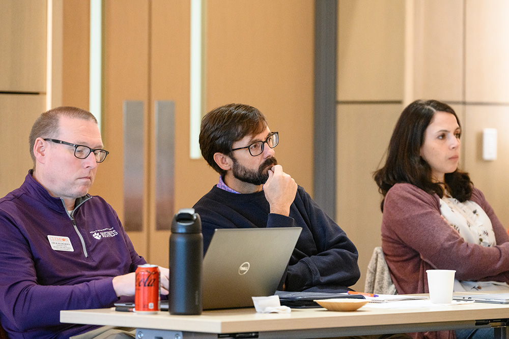 A group of three adults attentively listening during a presentation, with one person taking notes on a laptop.