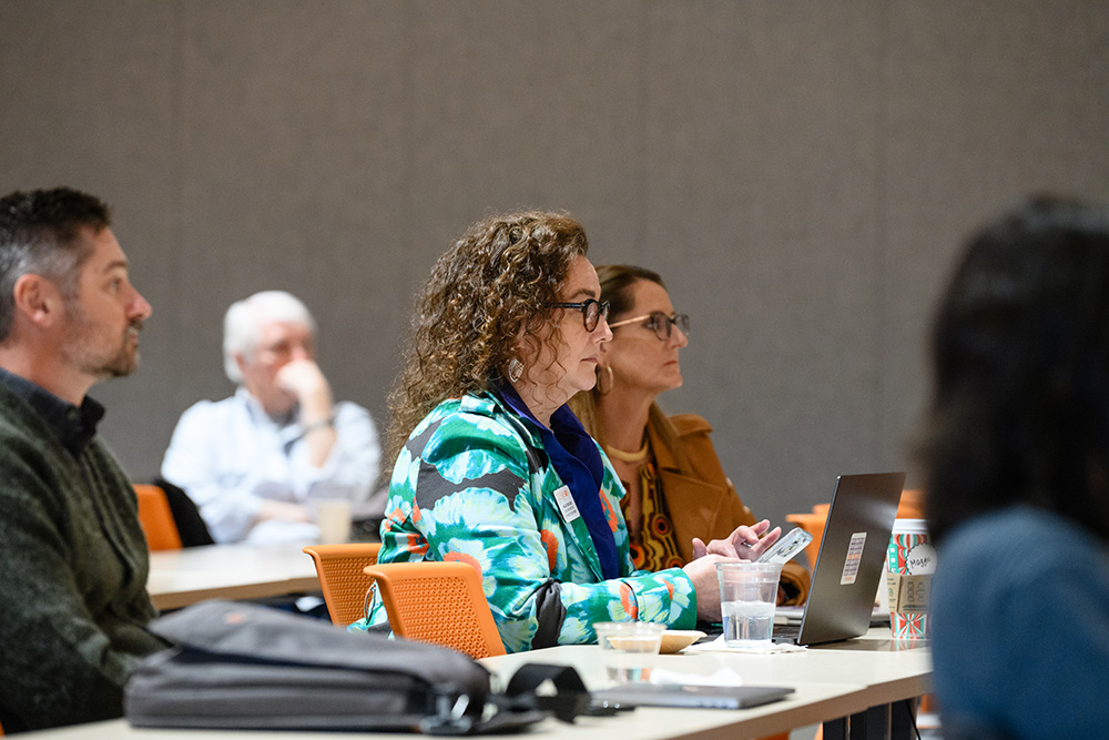 A woman with curly hair sits in a meeting, focused on her laptop while colleagues listen attentively in the background.