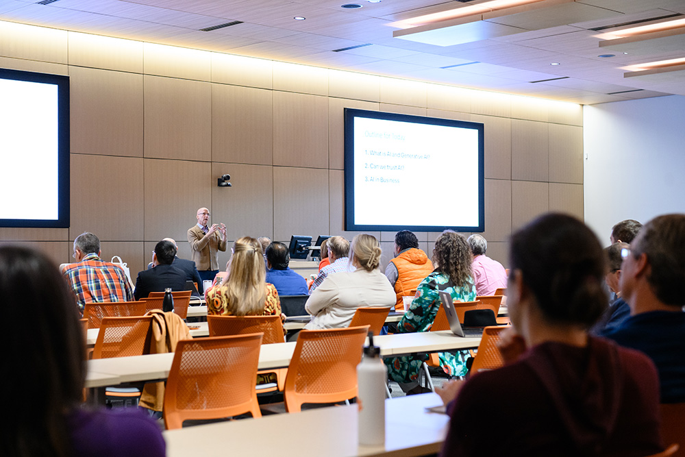 A presentation taking place in a seminar room, with an audience attentively listening to a speaker in front of a large screen.